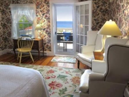 A cozy room with floral wallpaper, white armchairs, desk, and a view of the ocean through a glass door.