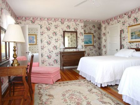 The image shows a floral-themed bedroom with two beds, a floral carpet, wooden furniture, and artwork on the walls, exuding a vintage feel.