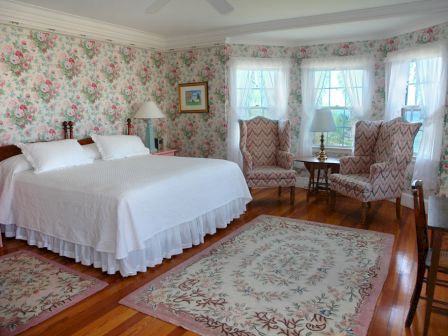 A cozy bedroom with floral wallpaper, a large bed with white bedding, two armchairs, rugs, and a lamp by the window on wooden floors.