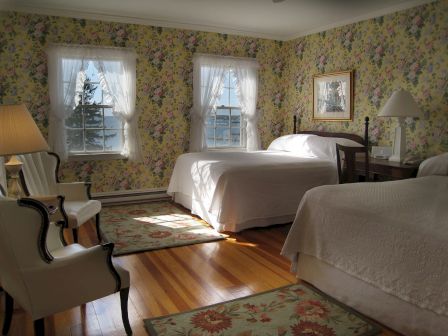 A cozy bedroom with two beds, floral wallpaper, wooden floor, and bright natural light from windows with sheer curtains.