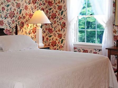 A cozy bedroom with floral wallpaper, a bed with white linens, framed art, side lamps, and a window with curtains.