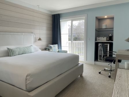 A cozy bedroom with a large bed, modern decor, and a small workspace, overlooking a bright balcony through a sliding glass door.