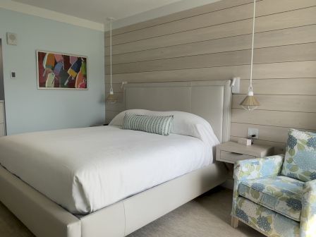 A modern bedroom with a large bed, wooden accent wall, floral armchair, and colorful framed art on a light blue wall.