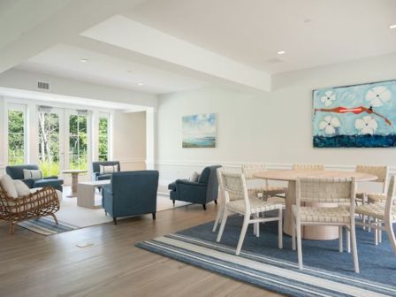 The image shows a room with blue armchairs, wicker chairs, a round table, and colorful paintings on the walls, with large windows.