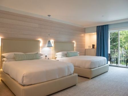 A modern bedroom with two double beds, blue curtains, and a balcony view. Warm lighting and minimal decor create a cozy atmosphere.