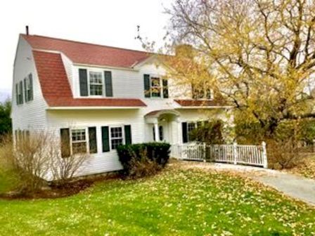 A two-story white house with red roof, surrounded by trees and a lawn with fallen leaves, is shown.
