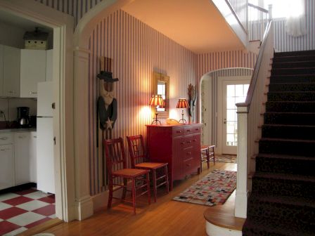 A hallway with striped walls, a red dresser, two chairs, lamps, and a staircase. There's a glimpse of a kitchen with checkered flooring.