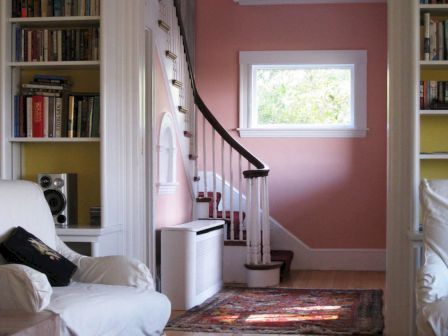 A cozy room with bookshelves, a couch, and a view to a staircase with a pink wall and window, showcasing a warm, inviting ambiance.