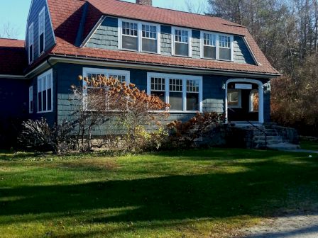 A two-story house with a red roof, large windows, and a small front porch, surrounded by a grassy yard and trees in the background.