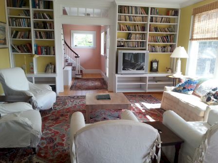 A cozy living room with white armchairs, bookshelves, a TV, rug, and a window with blinds in a sunlit setting.