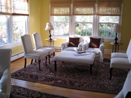 A cozy living room with white furniture, wooden floor, patterned rug, and sunlight streaming through windows with bamboo blinds.