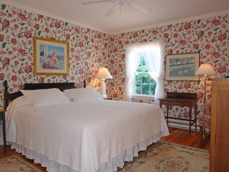A cozy bedroom with floral wallpaper, a large bed, paintings, lamps, and a window with sheer curtains above a small wooden desk.