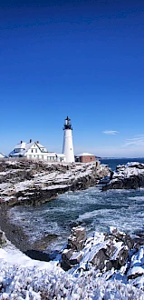 A snow-covered coastal scene with a lighthouse and building overlooking the ocean on a clear blue day.