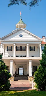 A white civic-style building with columns and an American flag on a spire, framed by green trees beneath a clear blue sky.